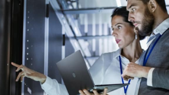 A man holding a laptop standing next to a woman pointing at a control panel